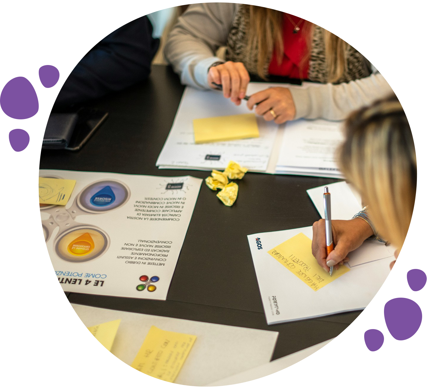 Three people are collaborating at a table with documents, sticky notes, and crumpled paper. The atmosphere is focused and analytical.