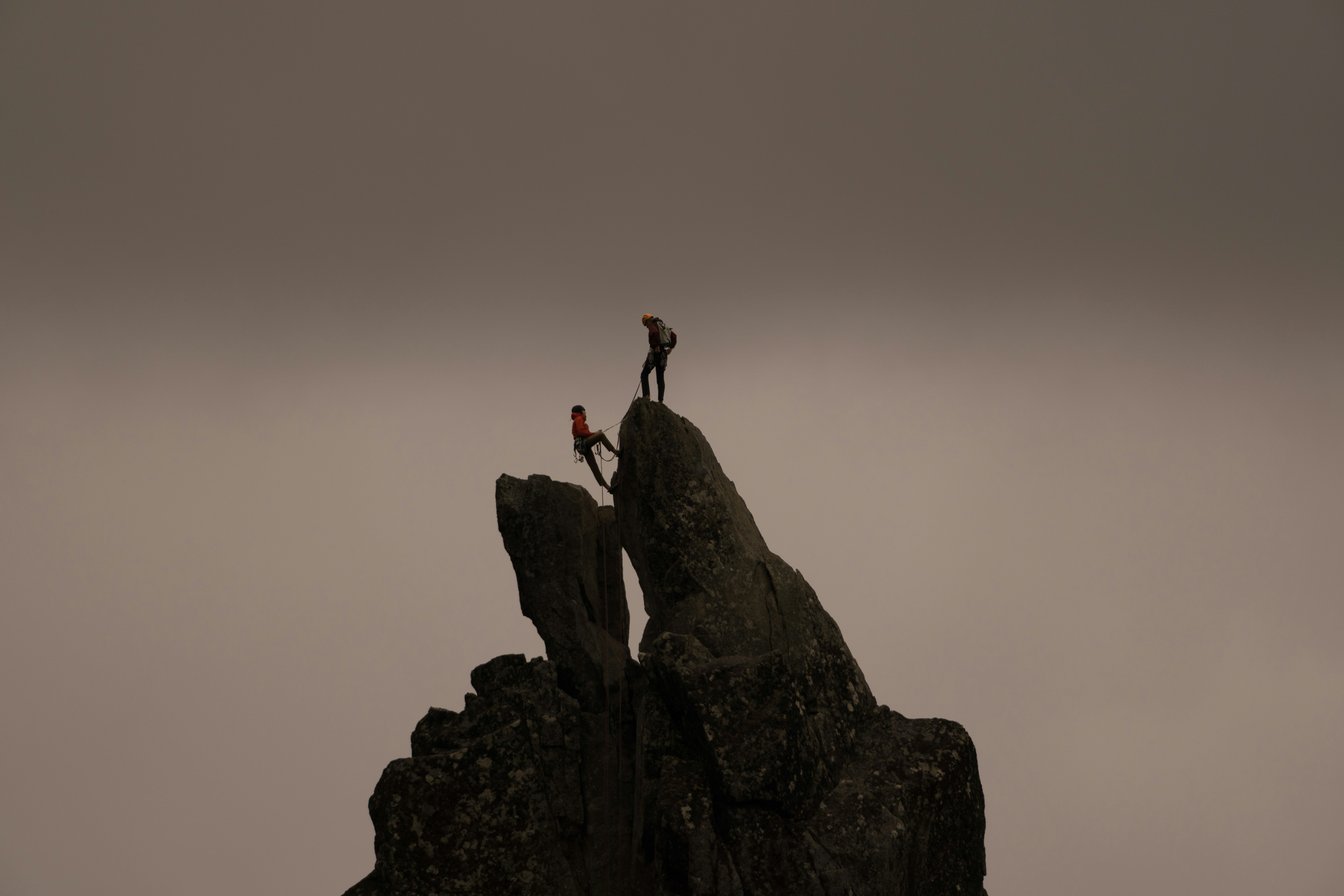 Two climbers atop steep jagged rocks, one standing and the other seated, against an overcast sky. The scene conveys adventure and triumph.