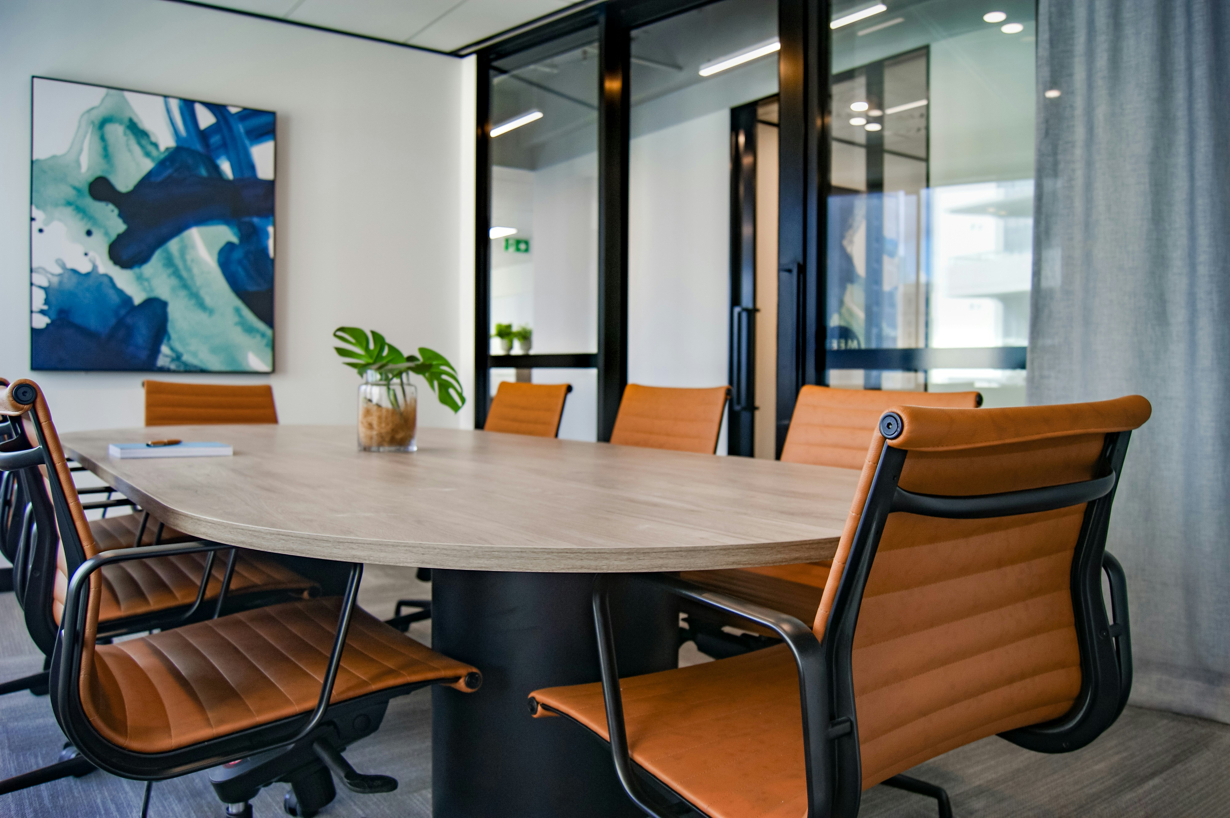 Modern conference room with an oval wooden table, surrounded by six orange chairs. A blue abstract painting and a plant in a glass jar add accents.